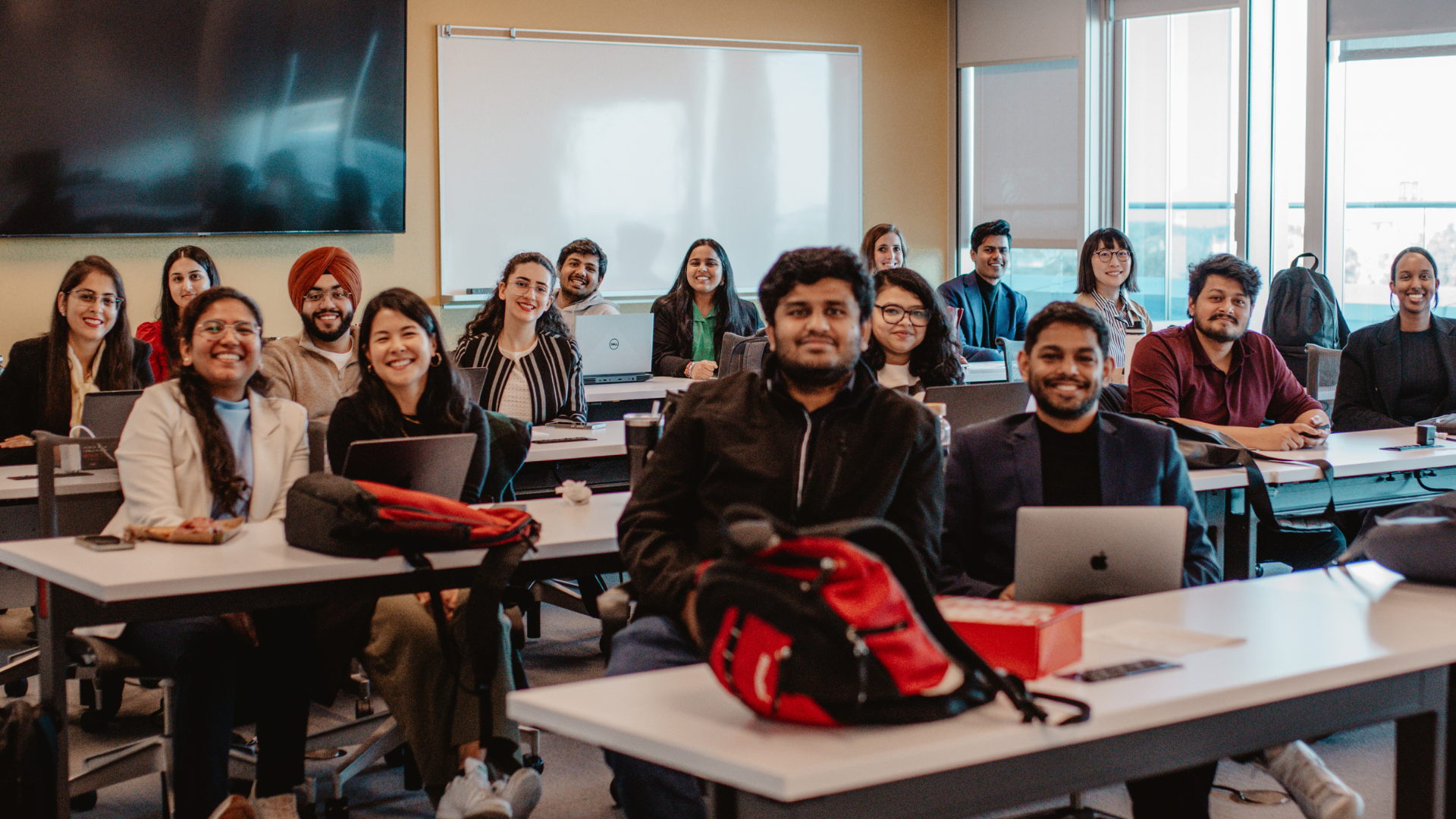 A diverse group of smiling students sits at desks in a classroom setting, some with laptops and bags.