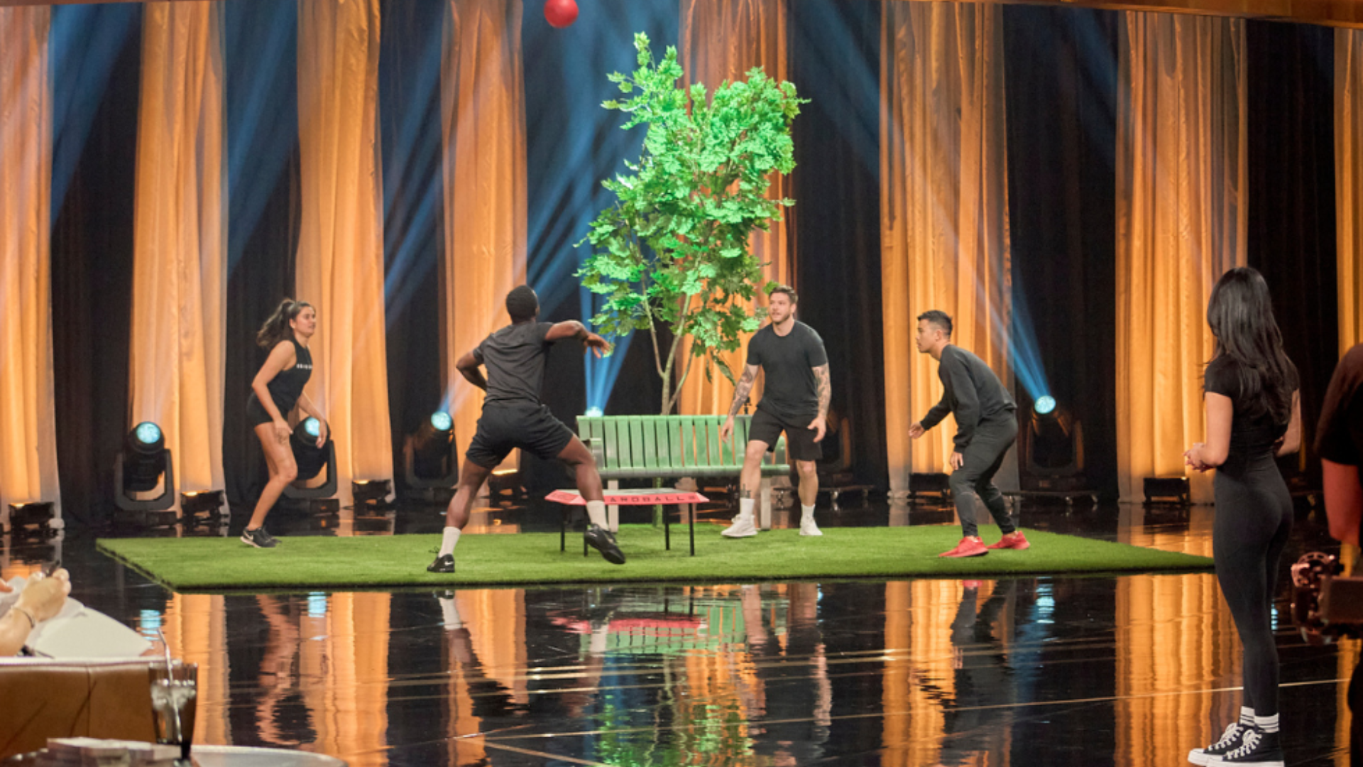 Four people play boardball on artificial grass in a studio as a man prepares to spike the airborne ball.