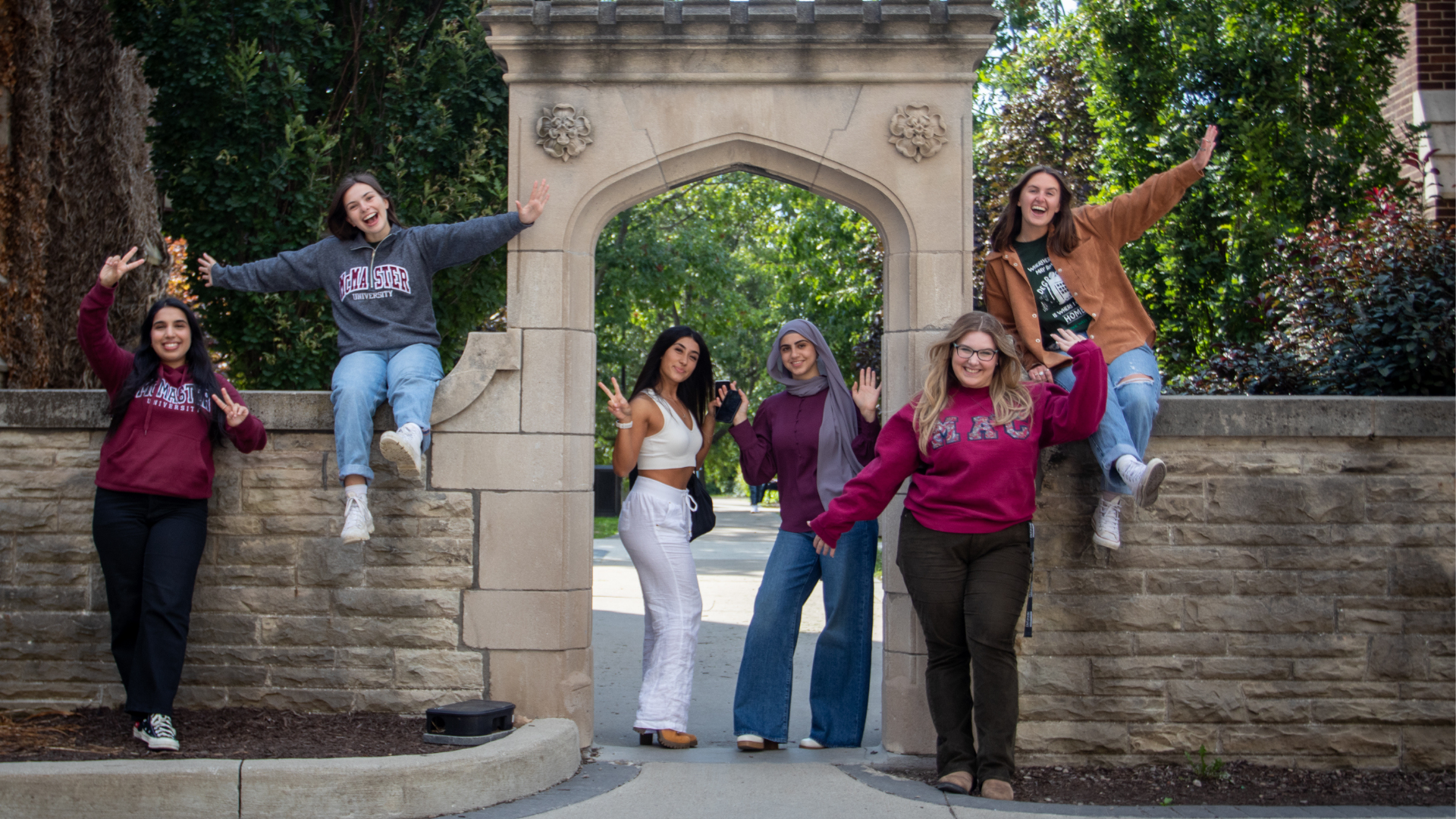 Six smiling female DeGroote students pose at the stone arch, some standing and sitting, waving and making peace signs.