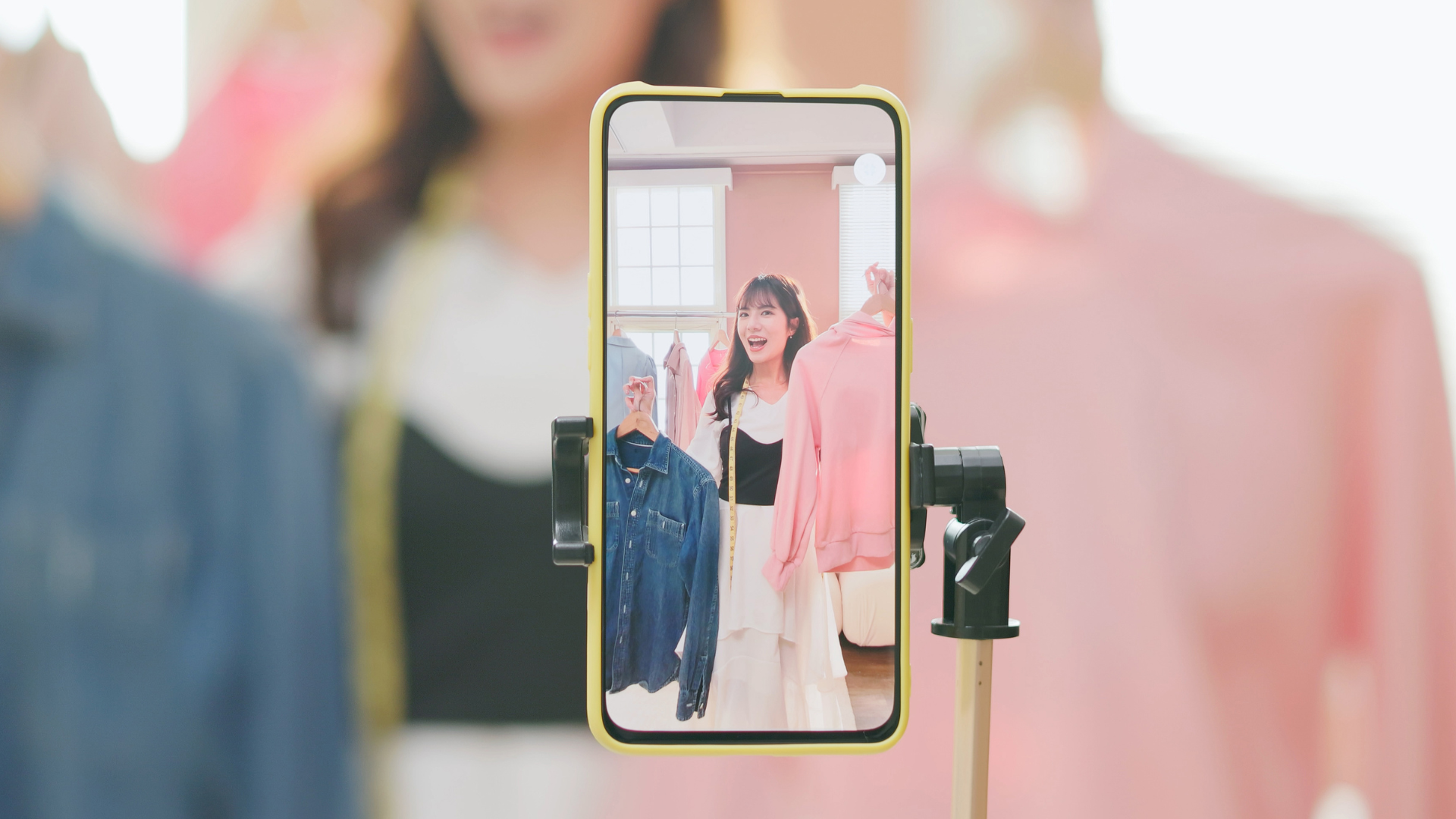 A cellphone recording a female influencer smiling as she holds up a pink blouse and jean jacket.