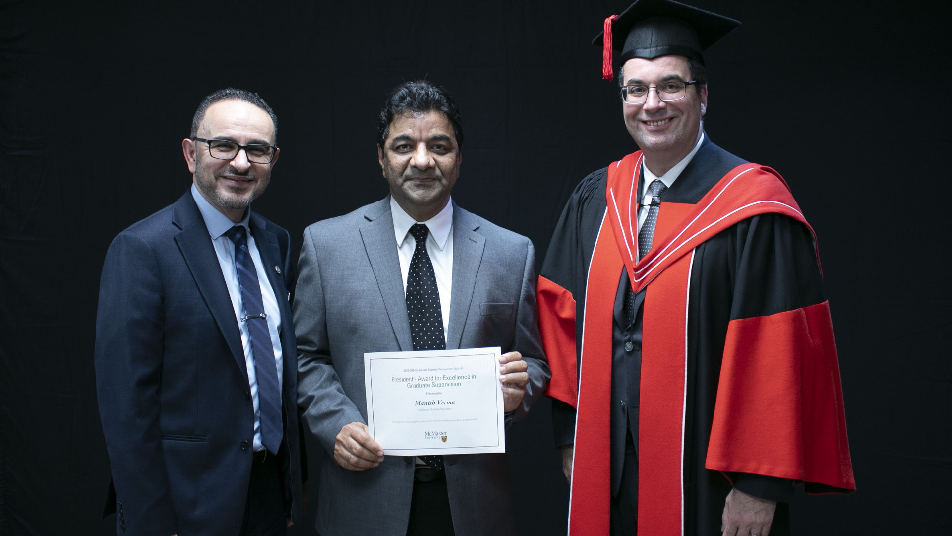 Three businessmen pose for a photo. One man holds a certificate, while the other man is wearing graduation attire.