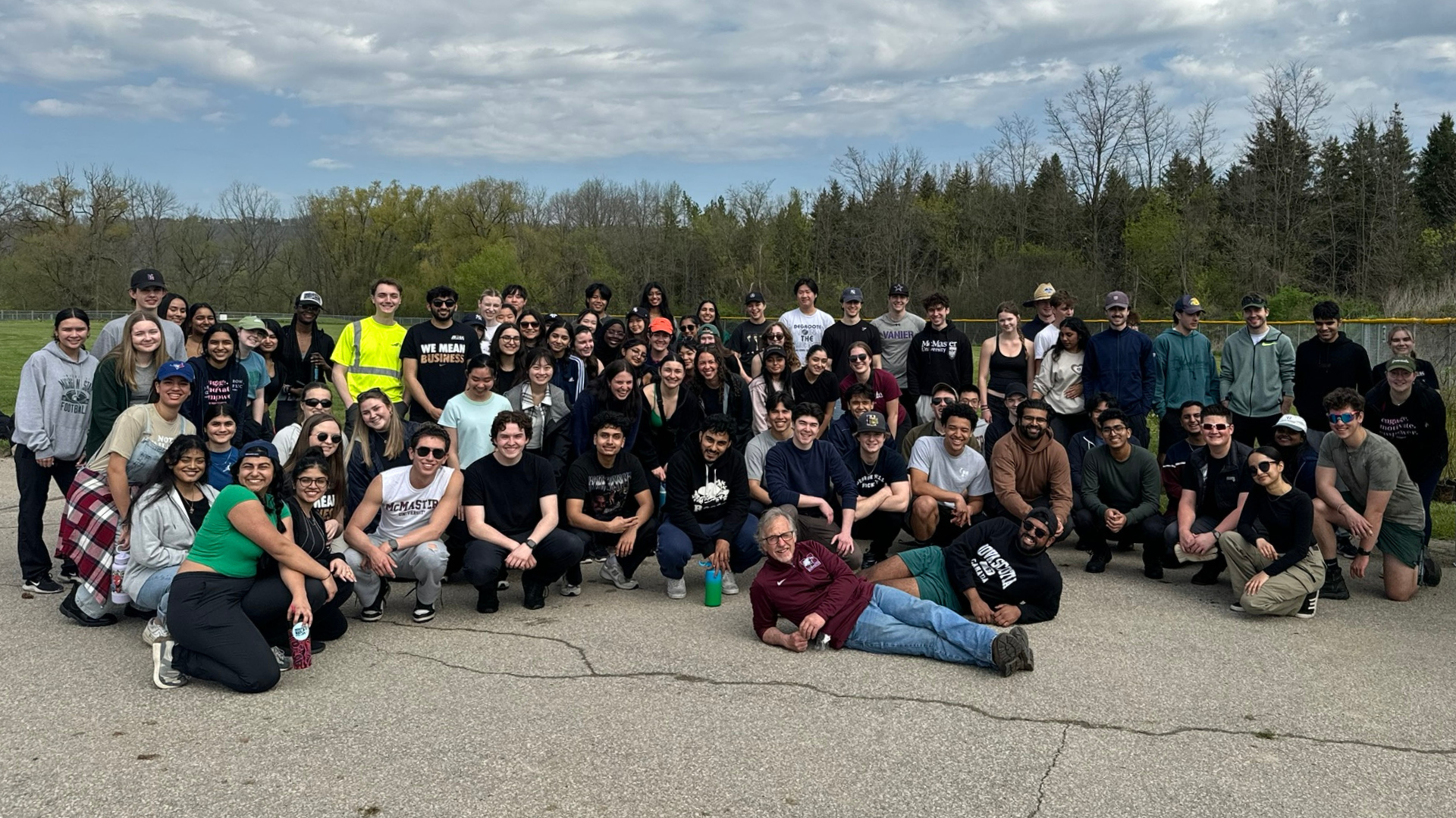 A large group of diverse students and a professor pose outdoors on a paved surface with a field and trees in the background.