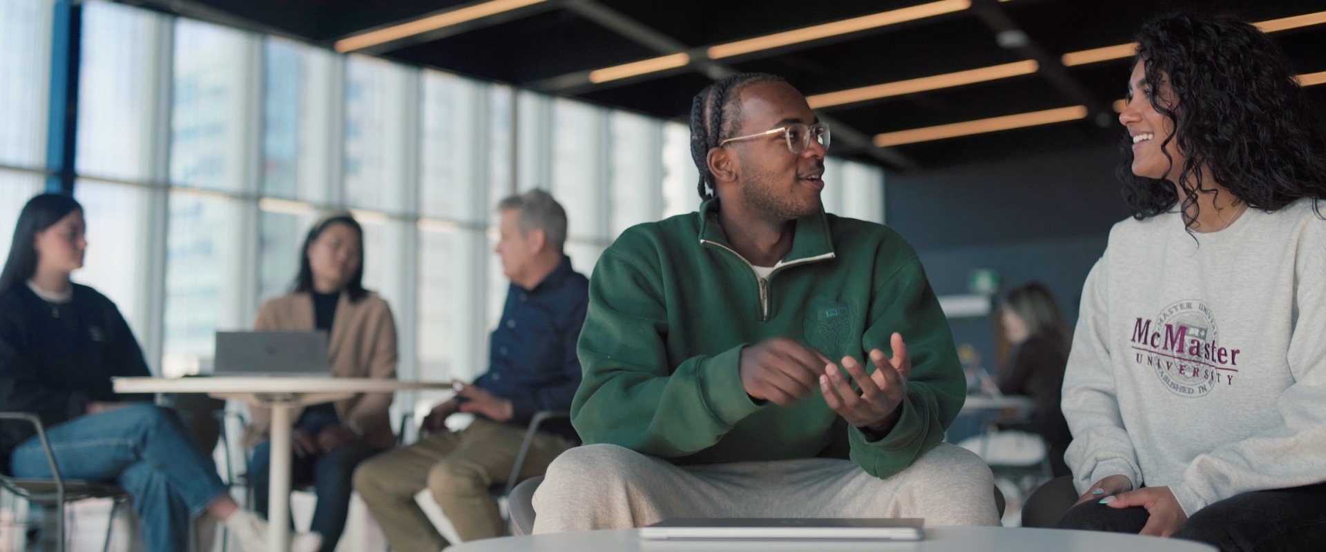 Two university students (a Black man with glasses and a Black woman) are sitting at a coffee table, smiling and talking.