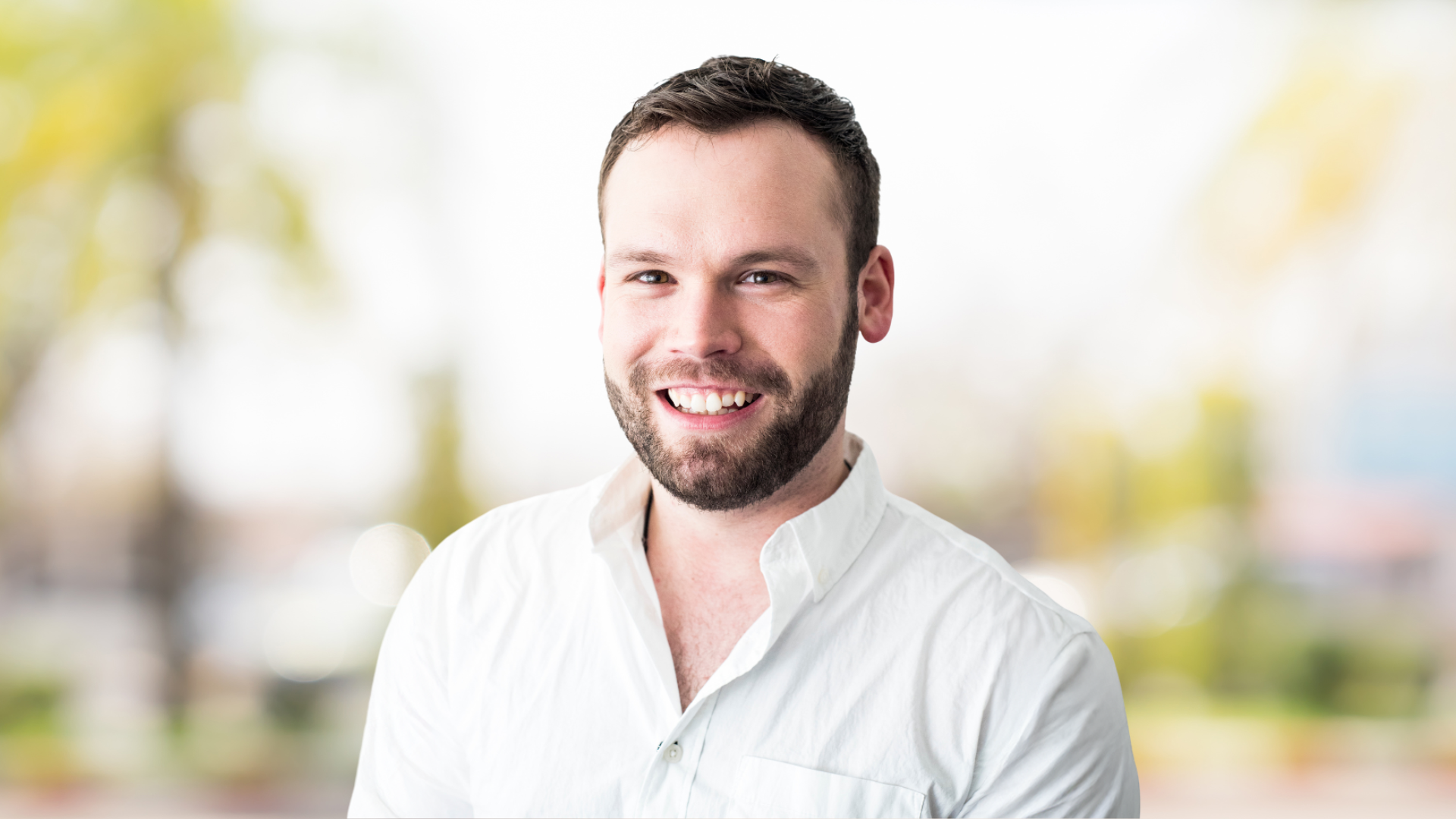 Person wearing a white collared shirt, posed in front of a bright outdoor background with soft greenery.