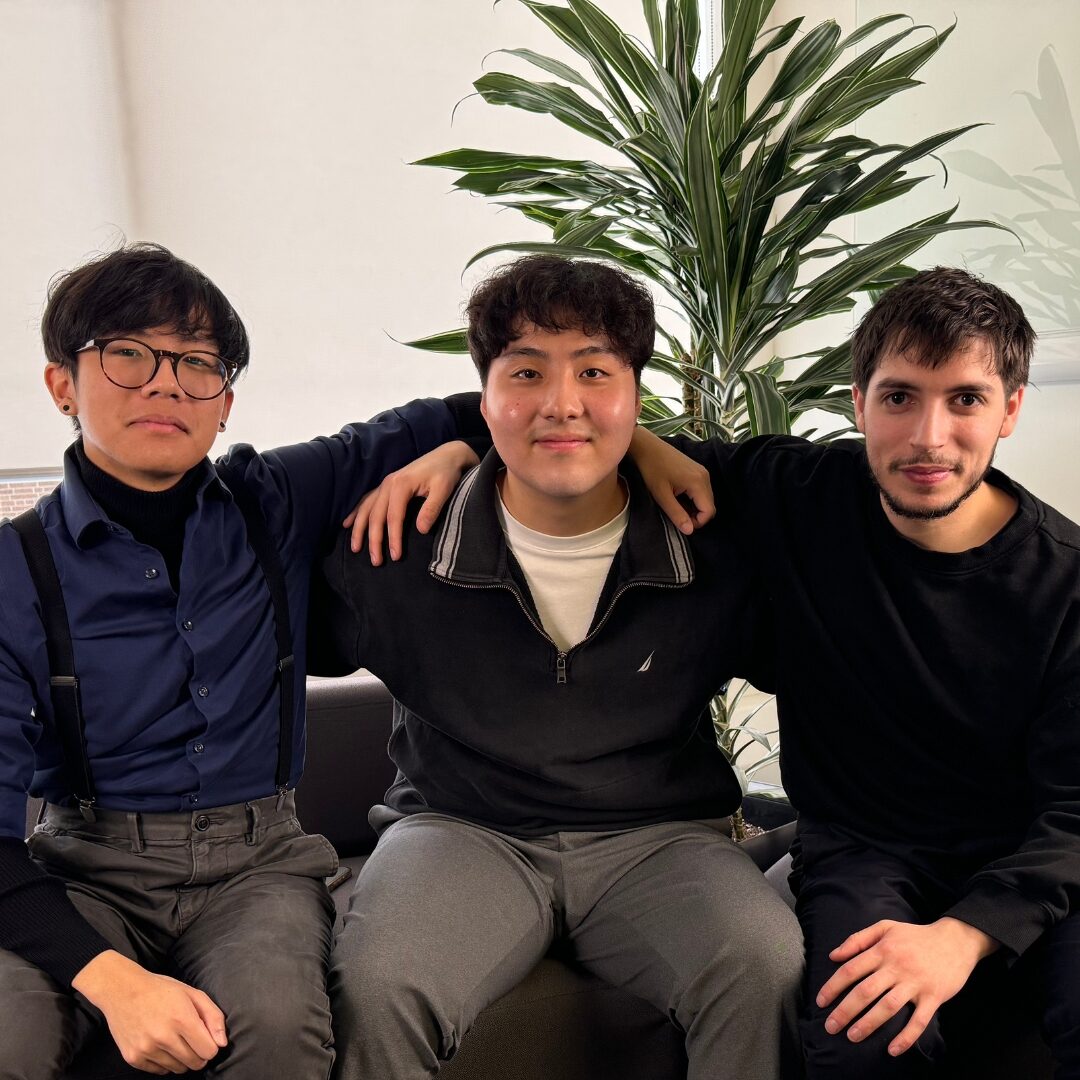 Three people sitting together on a couch with their arms around each other, with a plant behind them and a phone resting on the seat.