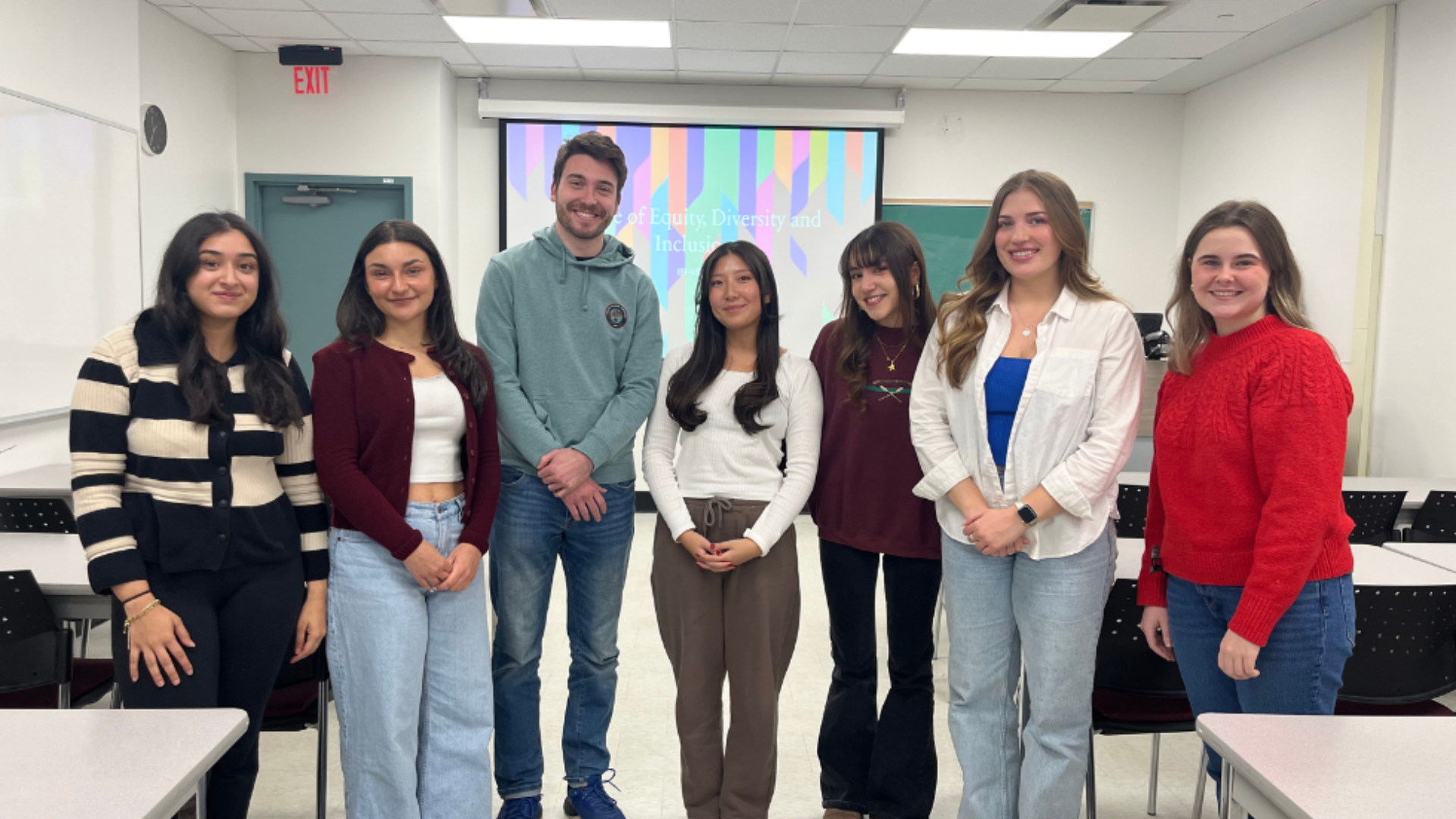 Group of students standing at the front of a classroom with a colorful presentation slide projected behind them.
