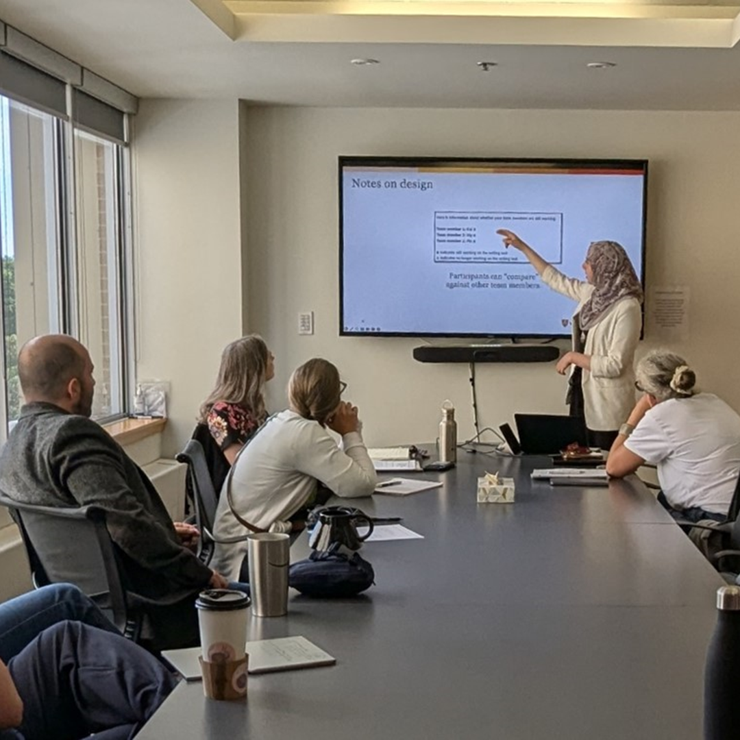 Person presenting at the front of a meeting room, pointing to a slide while attendees sit around a conference table.