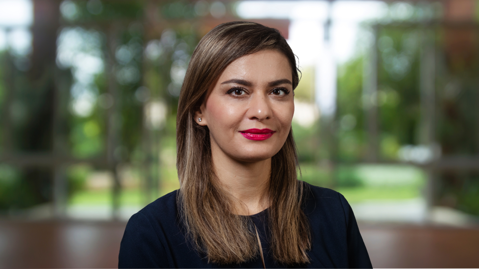 Person with long brown hair wearing a dark top, posed indoors with large windows and greenery in the background.