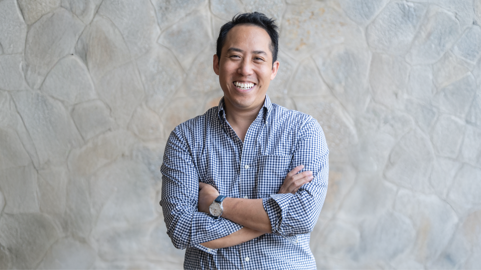 Person standing with arms crossed, wearing a checkered shirt against a textured stone wall.